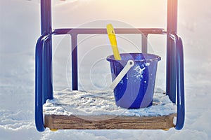 Blue plastic baby bucket with yellow paddle stands on wooden metal swing seat in winter on sunny day