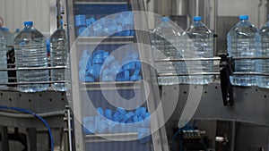 Blue plastic lids are lifted on a conveyor belt. Against the background, five-liter water bottles move along the production line.