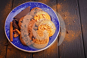 On a blue plate are biscuits, black chocolate and a few cinnamon sticks. On a wooden table. Inside. View from above.