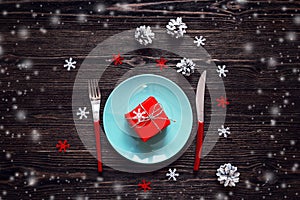 Blue plate with gift box, red knife and fork, and christmas decorations on dark wooden table. Christmas table setting.