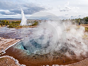 Blue pool with vapour in Haukadalur geothermal area with Strokkur big geyser steam in the background. One of the most visited and