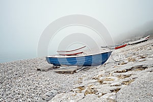 Blue and red fishing boats on a rocky pebble beach in the early morning with foggy mist on background