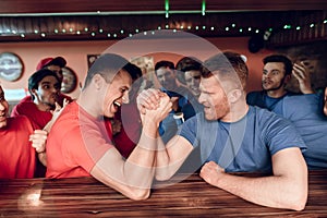 Blue and red team fans arm wrestling at sports bar with fans in background.