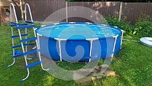 A blue round above-ground frame pool stands on a grassy lawn. A ladder stands next to it. The pool is made of PVC. Behind the lawn