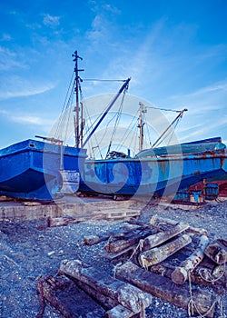 Blue rusting fishing boats on the seashore at the setting sun. Pebbles and wooden support logs in the foreground and a blue sky