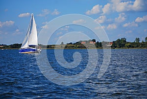 A blue sailboat sails on the lake. The shore and some houses in the background. Yacht sailing in Masuria (Mazury).