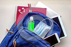 A blue school bag pack with sanitizer and a face mask, pencils earphones, tablet, computer and a smartphone. Back to school during