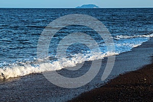 Blue sea over volcanic sand beach. Distant island view. Relaxing sea wave surf over seashore.