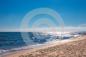 Blue sea sparkling in the sun with a wave, sandy beach and blue sky on a summer day