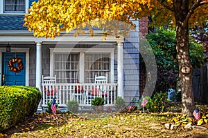 Blue shinngled two stor house entrance with porch and rocking chairs and American flags and a fall wreath under a yellow-leafed