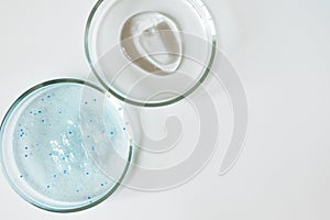 Blue shower gel with scrub grain with bubbles and transparent shampoo smudge in glass petri dish on white background