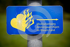 Blue sign in German: Ammergau Alps meditation trail. With a yellow flaming heart