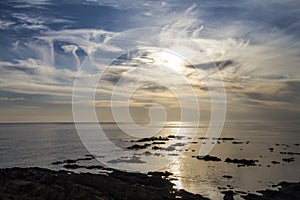 Blue sky at dawn covered by strange cloud formations on a flat sea with black rocks that stand out in the reflection of the sun on