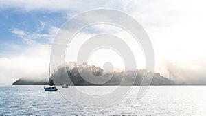 Blue sky, white clouds, mist & aquamarine seas at Akaroa Harbor. Anchored are fishing boats & sailboats, along the Port at Akaroa