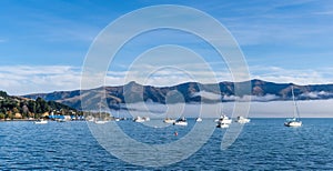 Blue sky, white clouds, mist & aquamarine seas at Akaroa Harbor. Anchored are fishing boats & sailboats, along the Port at Akaroa