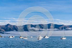 Blue sky, white clouds, mist & aquamarine seas at Akaroa Harbor. Anchored are fishing boats & sailboats, along the Port at Akaroa