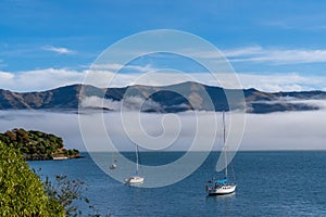 Blue sky, white clouds, mist & aquamarine seas at Akaroa Harbor. Anchored are fishing boats & sailboats, along the Port at Akaroa