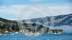 Blue sky, white clouds, mist & aquamarine seas at Akaroa Harbor. Anchored are fishing boats & sailboats, along the Port at Akaroa