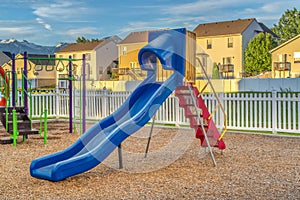 Blue slide with red stairs at a playground against homes mountain and blue sky