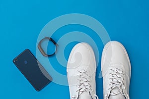Blue smart bracelet, mobile blue and white sneakers with a blue background. Sports style. Flat lay.