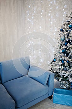 Blue sofa near Christmas tree, gifts on gray wall with light bulbs garlands background. New Year`s interior. copy space