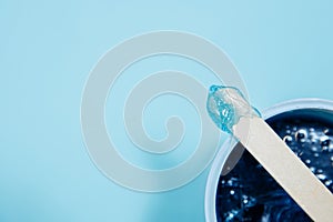 Blue soft wax lies on a depilation spatula. Sharpness is on wax with a spatula. A wooden spatula lies on a jar of wax.