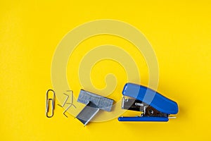 Blue stapler with metal staples and paper clip on yellow background. Top view. Copy, empty space for text. Minimalistic concept