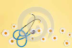 Blue stethoscope with chamomile flowers on yellow background. Top view with copy space