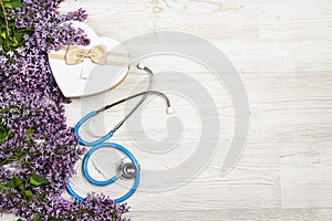 Blue stethoscope with lilac flowers and gift box on light wooden background. Top view with copy space. National Doctor`s day