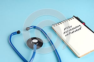 Blue stethoscope and a notebook with the inscription `Flu season ahead` on blue background.