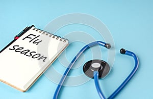 Blue stethoscope and a notebook with the inscription `Flu season` on blue background.