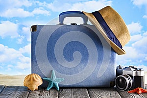 Blue suitcase for summer holidays, hipster hat; retro photo camera and shells on wooden table and beach