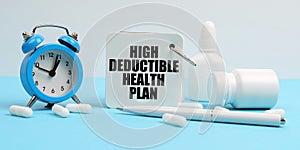 On a blue surface lie pills, an alarm clock and a notebook with the inscription - High Deductible Health Plan