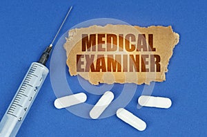 On a blue surface lie a syringe, pills and a cardboard sign with the inscription - MEDICAL EXAMINER