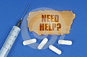 On a blue surface lie a syringe, pills and a cardboard sign with the inscription - Need help