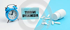 On a blue surface are pills, on a white surface an alarm clock and a plate with the inscription - Tardive Dyskinesia