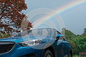 Blue SUV car covered with raindrops against green forest and rainbow sky. Car parked at outdoor parking lot. Road trip travel.