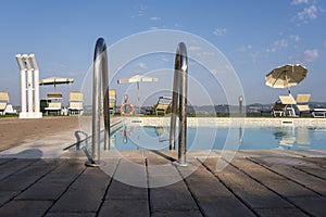 Blue swimming pool with beach lounger on wooden flooring with parasol, sun deck on sea view for summer vacation