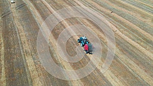 A blue tractor with a red baler collects straw into bales.