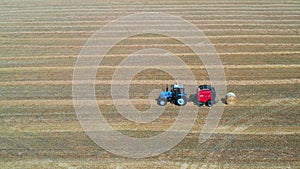 A blue tractor with a red baler collects straw into bales.