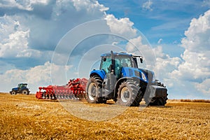 blue tractor with red harrow in the field against a cloudy sky, agricultural machinery work