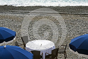 Blue umbrellas, reserved tables with white tablecloths on the pebble beach of the Promenade des Anglais in Nice, France, await gue