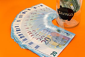 Blue used banknotes of twenty euros stacked in fans on an orange background and a transparent glass piggy bank .