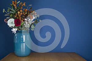 Blue vase on wooden table with plastic fake flower bouquet near blue wall, modern stylish decoration in living room with