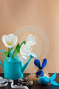 Blue watering can with white tulips and two blue Easter eggs in form of rabbit on brown background.