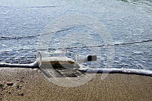 The blue wave of the sea breaking on the abandoned empty bottle and the reflection of the glass on the shoreline