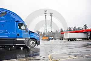 Blue wet big rig industrial semi truck driving on the truck stop parking lot with fuel station at winter snow and rain weather