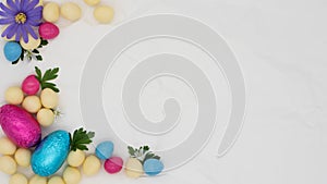 Blue and white flowers green leaves and candy pink wrapped Easter eggs on a scrunched plain white paper background with copy space