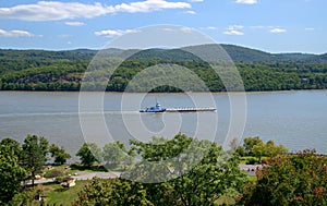 Blue and white tug boat was seen pushing a long barge down the Hudson River. There are small green tree covered hills in the.