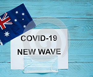 On a blue wooden background is a sheet with the inscription: covid -19 new wave, a medical mask and an Australian flag
